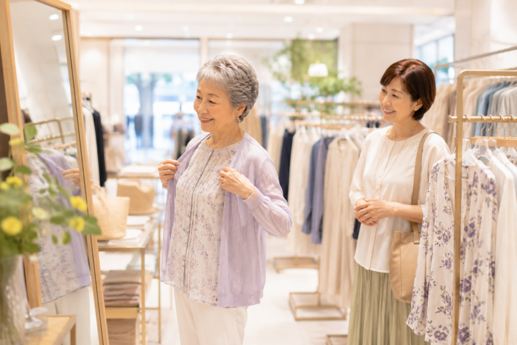 ショッピングモールの洋服店で鏡を見ながら自分で洋服を選ぶ高齢女性と同行スタッフ